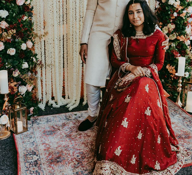 Bride & groom sit in front of floral archway complete with Moroccan styled rug and pillar candles 