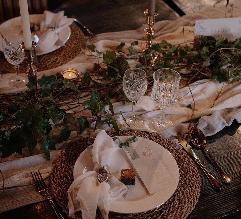 Real foliage table runner with a wicker placemat, white plate, white and grey stationery, mini brownie favour, and white napkin with pink ribbon