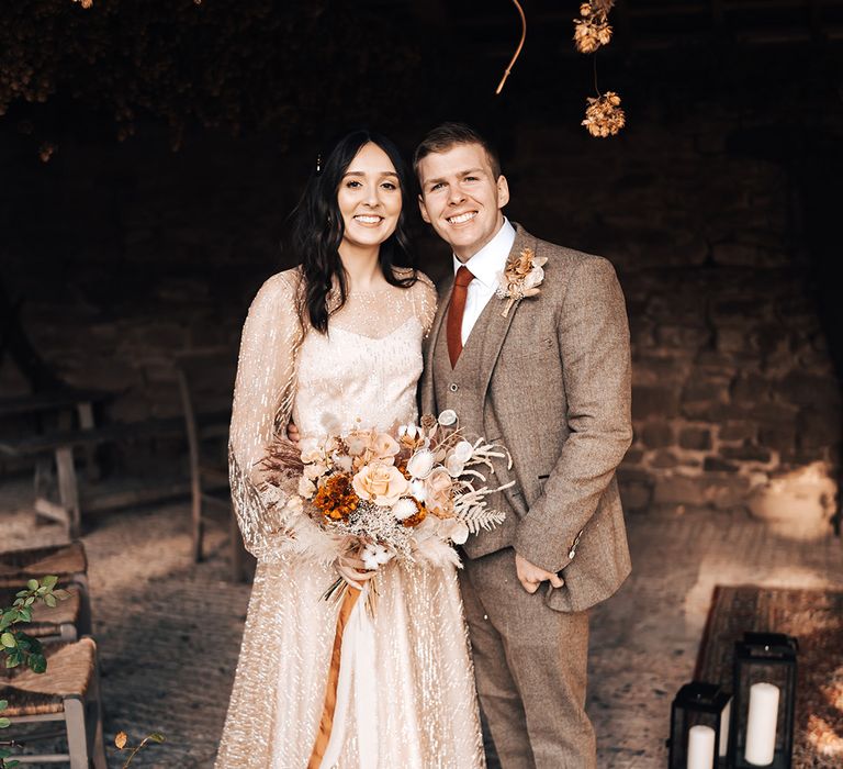 Bride in gold wedding dress stands with her groom in three piece brown suit with burnt orange tie