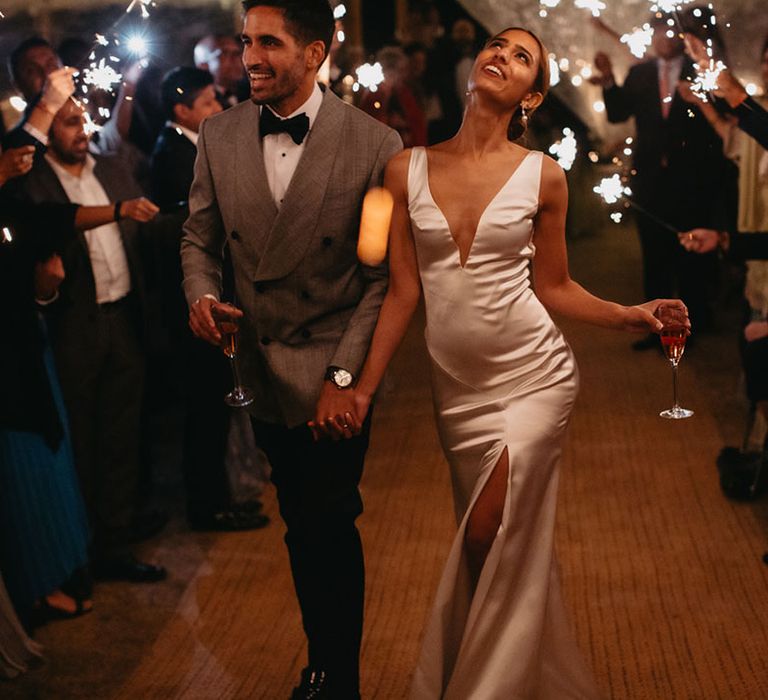 Bride wears v-shaped silk fitted wedding dress and walks with her groom in black-tie through sparkler exit 