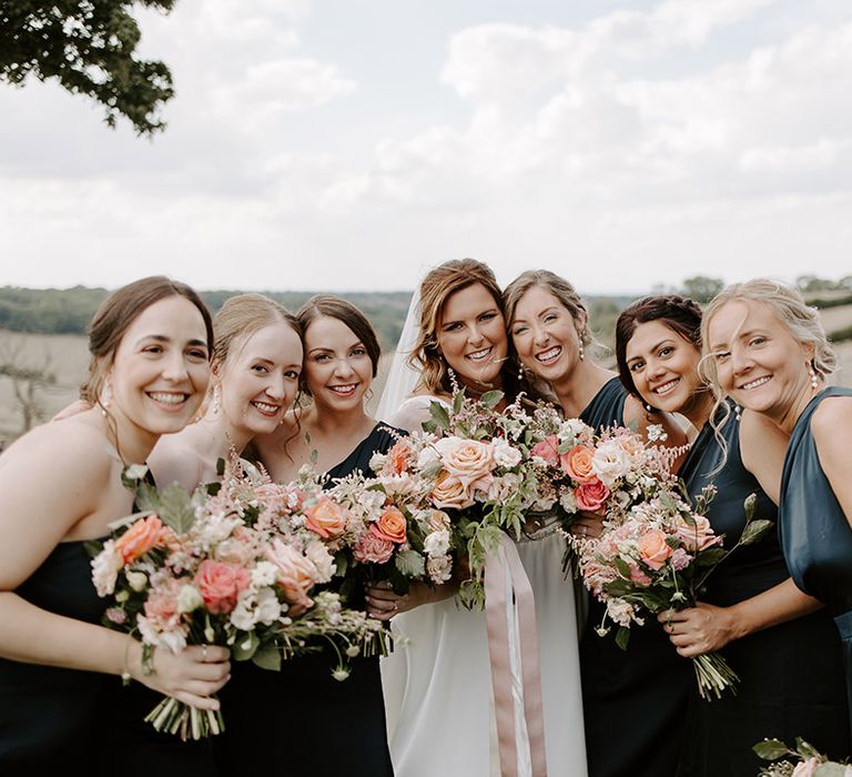 Bride poses with her bridesmaids wearing one shoulder green satin bridesmaid dresses and holding pastel bouquets 