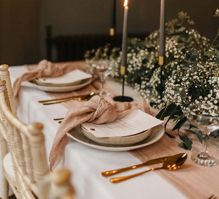 Wedding menu with neutral pink napkin, rose gold cutlery and sage green ceramic plates in front of foliage table decorations and sage green tall candles