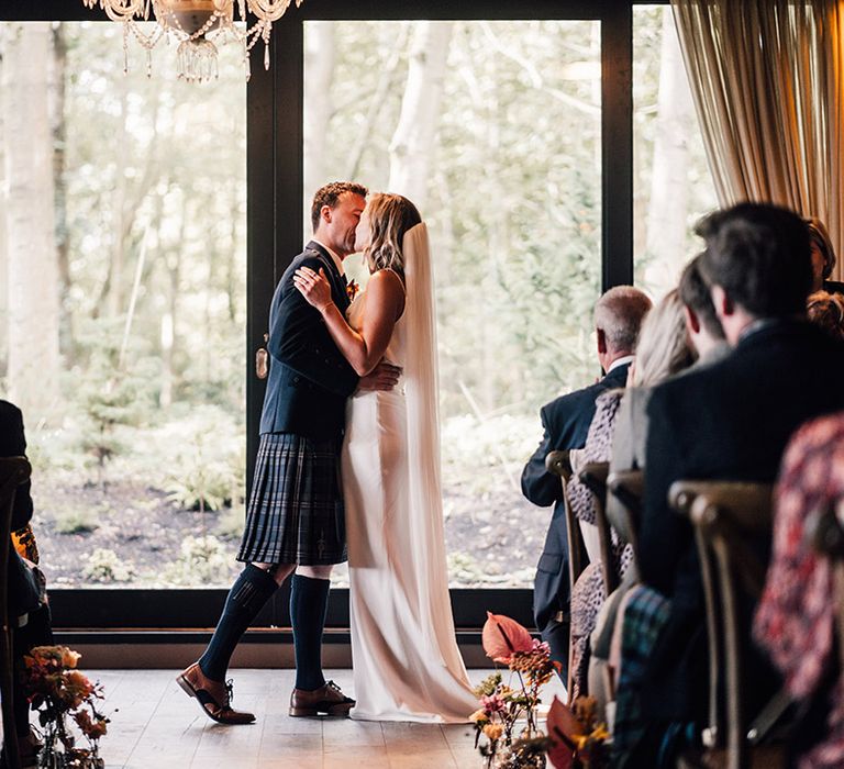 Bride wearing cathedral veil kisses her groom in front of large window