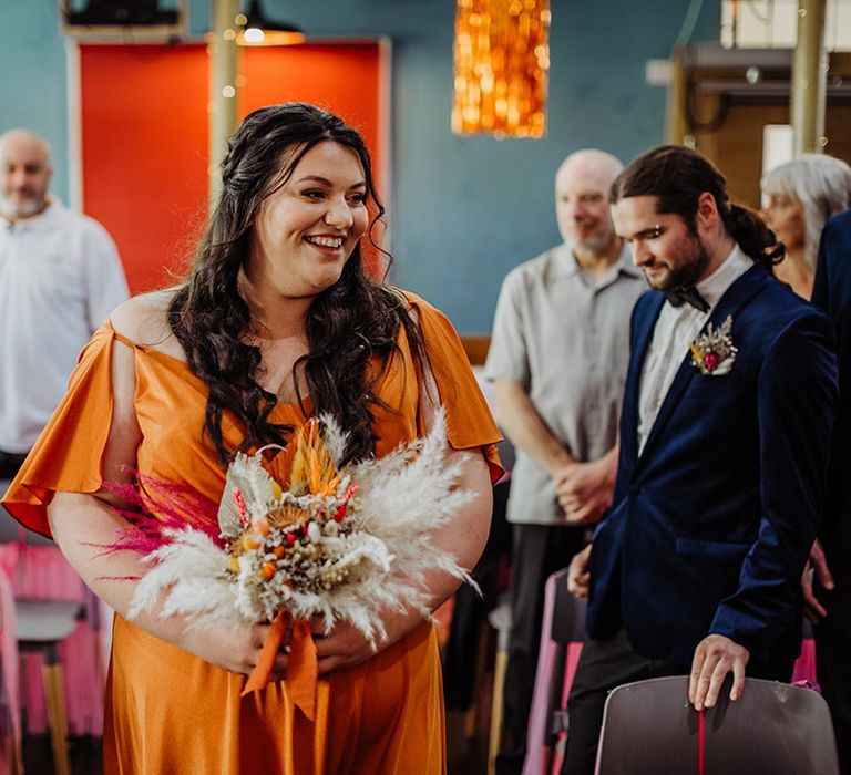 Bridesmaid walking down the aisle wearing off the shoulder orange bridesmaid dress with short flute sleeves and an autumnal floral bouquet with pampas grass and orange ribbon