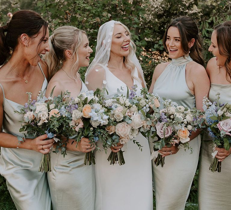 Bride in flower applique veil laughs with the bridesmaids in sage green dresses holding pastel bouquets 