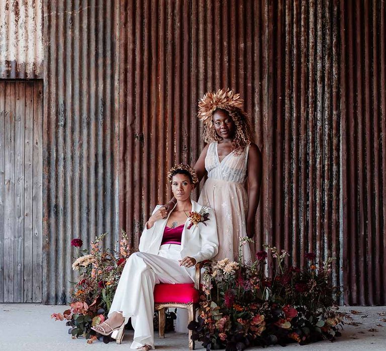 Bride in sleeveless floaty peach gown with embroidered stairs and gold leaf crown standing with bride sitting on velvet and gold chair wearing white silk suit with autumnal dried floral boutonniere and gold bridal crown 