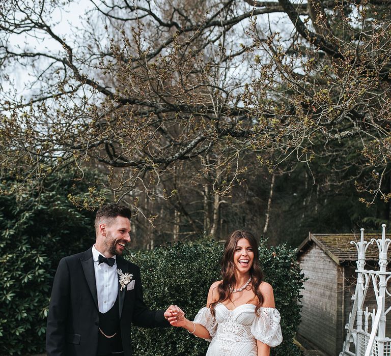 Groom in three piece black tux holding hands with the bride in a boho fitted lace dress for their wedding 