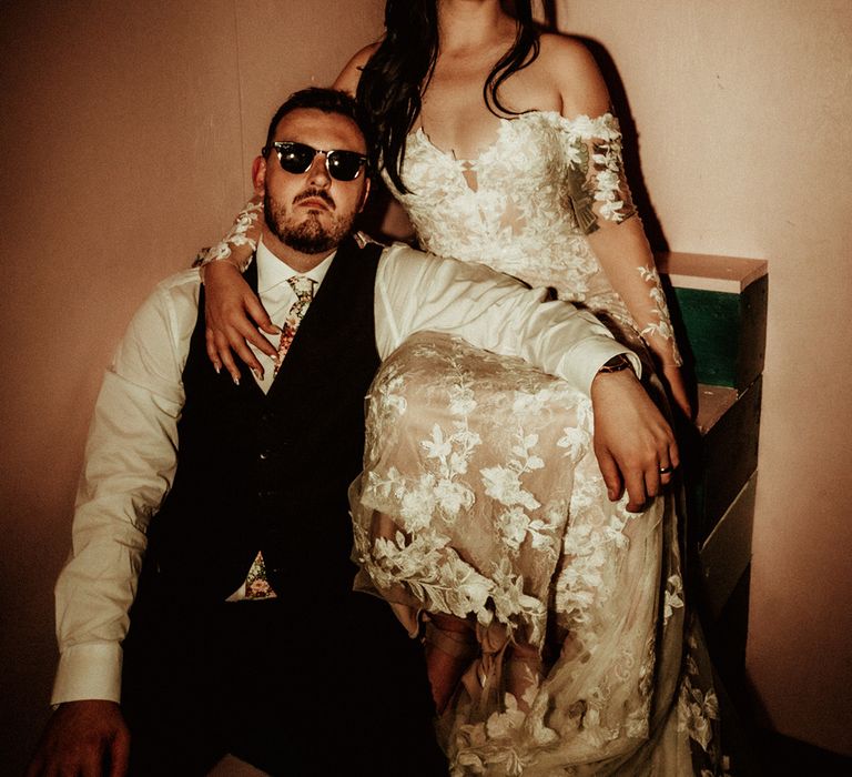 Bride in off the shoulder bridal gown with long, sheer sleeve and lace detailing wearing cat eye bejewelled sunglasses posing with groom in black waistcoat and flower patterned tie wearing Raybans sunglasses