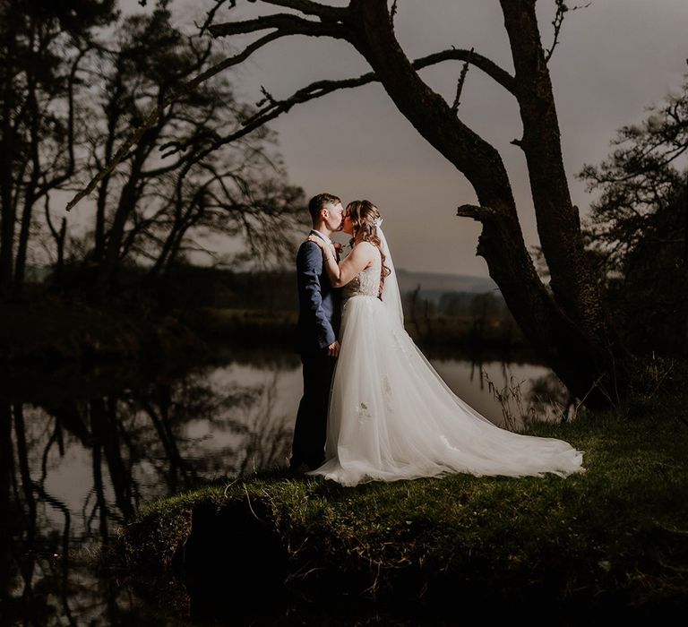 Bride in lace short sleeve wedding dress kisses her groom at night as their reflection can be seen in the water 