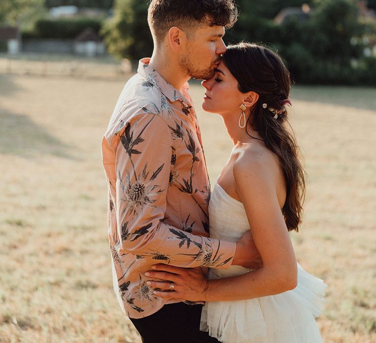 Groom in pink summer Ted Baker shirt kissing the bride on the forehead in a strapless gown with ruffle detail in half up half down style