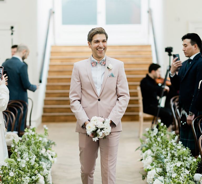 Man walks down the aisle in pastel pink suit and holds floral bouquet 