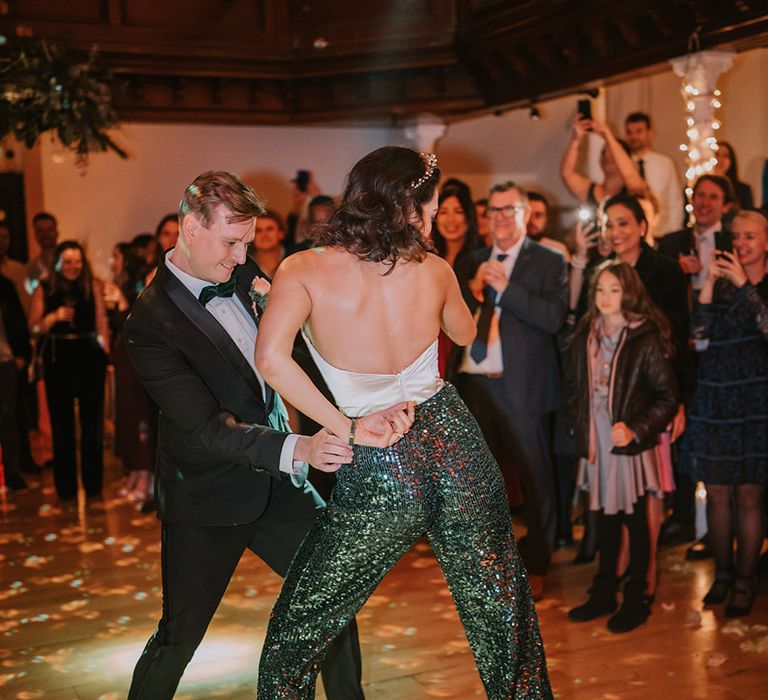 Bride in low-back detail taffeta crop top, dark green sequin trousers, chunky velvet green platform heels and bridal crown dancing with groom in classic black tux 