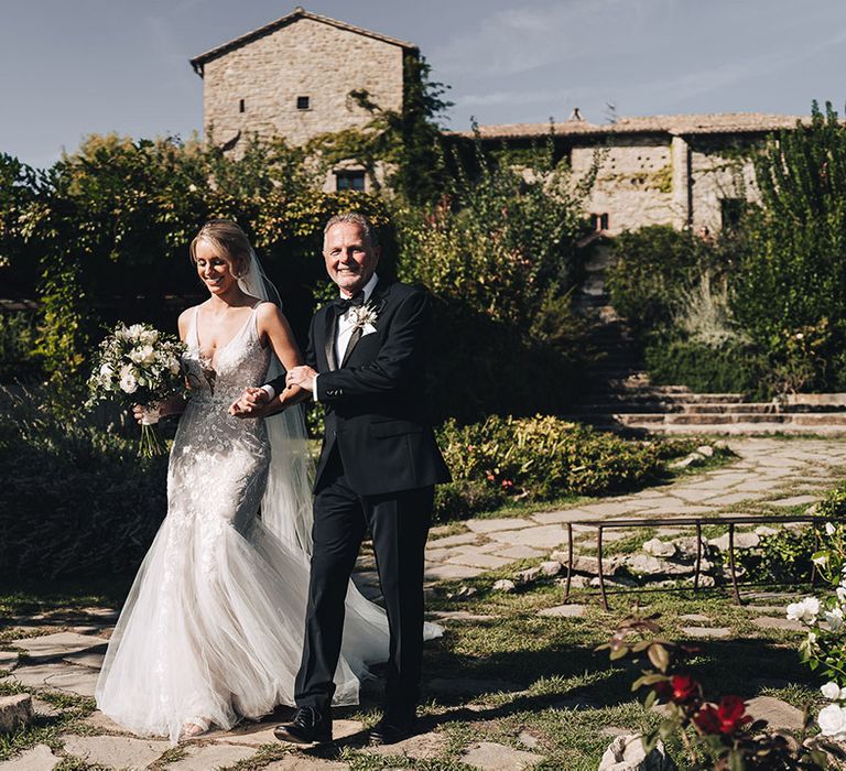 Bride in lace trumpet skirt wedding dress walks alongside her father in black-tie  