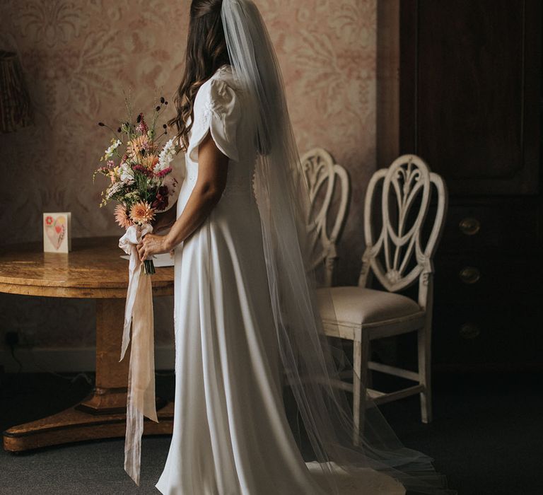 Bride with brown hair in a half up half down hairstyle with veil wearing a button back short puff sleeve wedding dress with rustic wedding flowers bouquet