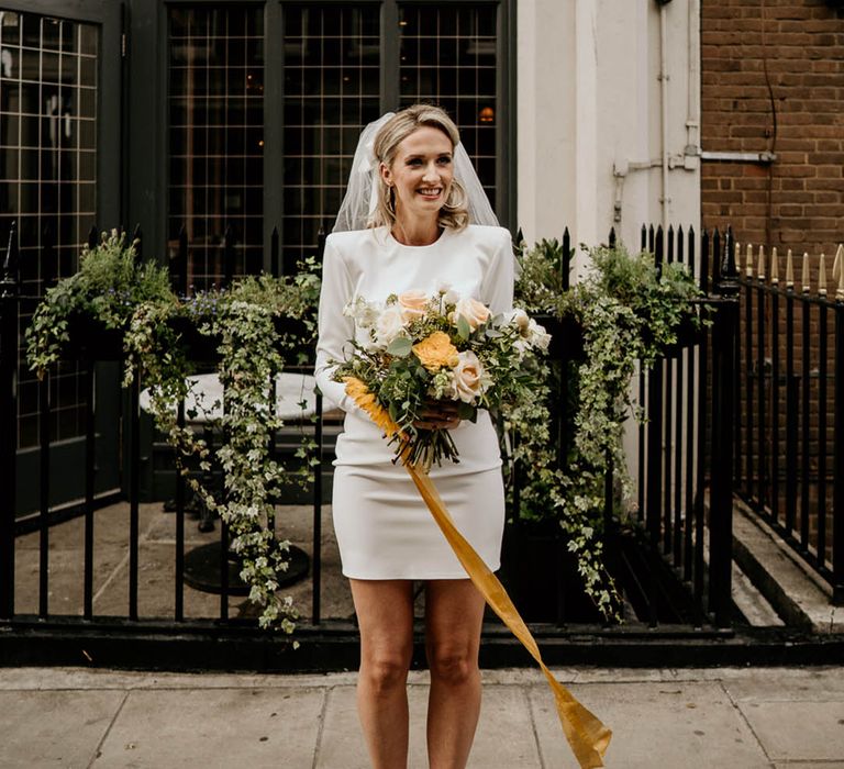 Bride in a short wedding dress with long sleeves and shoulder pads and yellow Manolo Blahnik pumps holding a sunflower bouquet 