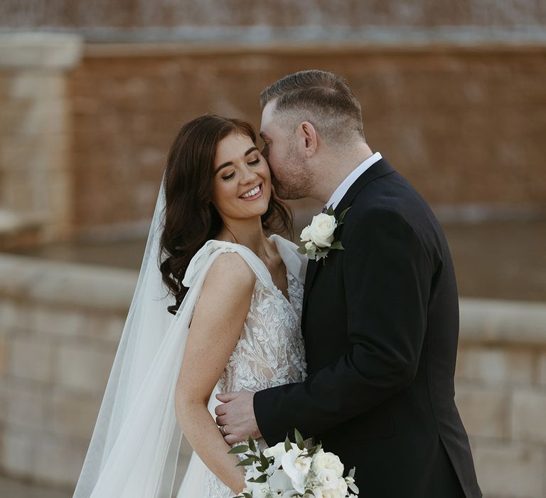 Bride holding a cascading wedding bouquet with all white wedding flowers including roses and orchids in a contemporary wedding dress posing with the groom for couple portraits