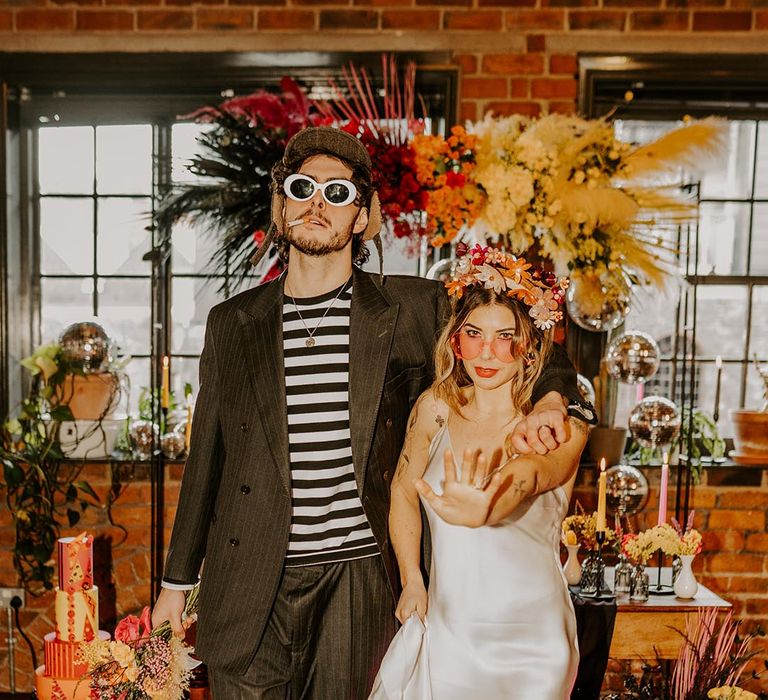 Bride in satin v-neck wedding dress, flower crown and open-toe pink fluffy heels and groom in dark pinstripe suit with black and white long sleeve t shirt, large white sunglasses and dark cap posing at 90s inspired grunge glam wedding shoot 