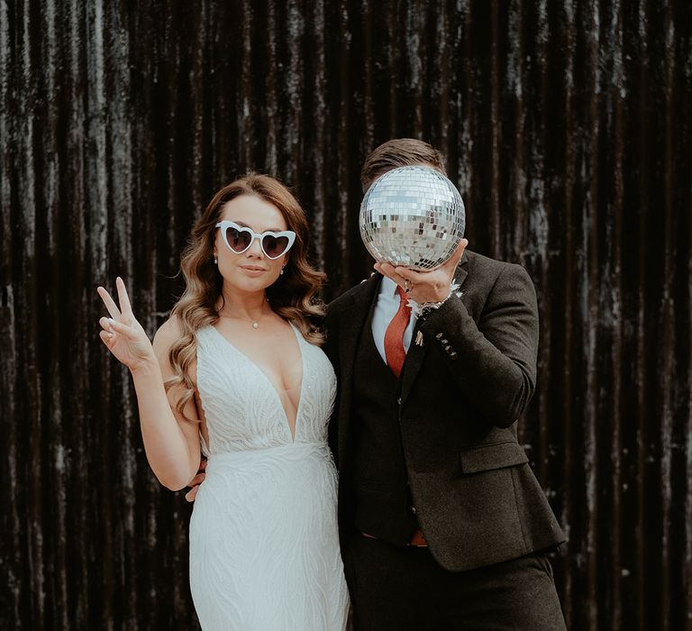 Bride in sequin patterned Made With Love wedding dress with white retro wedding sunglasses with the groom in dark suit holding disco ball decor over his face 