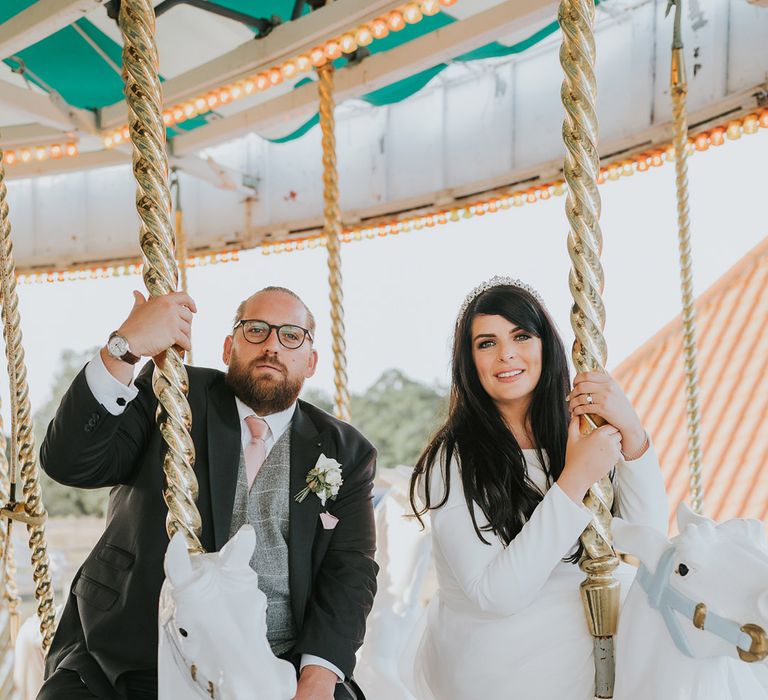 The bride in a high neck long sleeve wedding dress with tiara rides a horse on the carousel with the groom in a dark grey suit jacket 