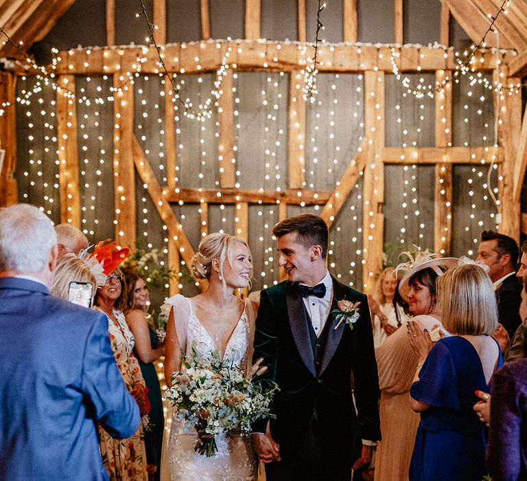 The groom in velvet tuxedo walks back down the aisle with the bride in a fitted lace wedding dress as a married couple 