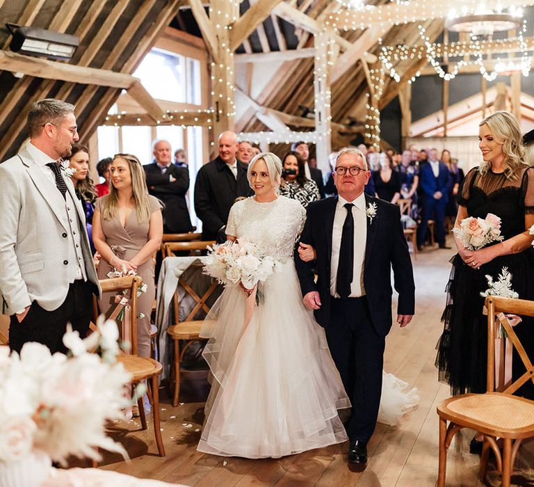 The father of the bride in black and white suit walks the bride down the aisle to the groom for the rustic barn wedding 