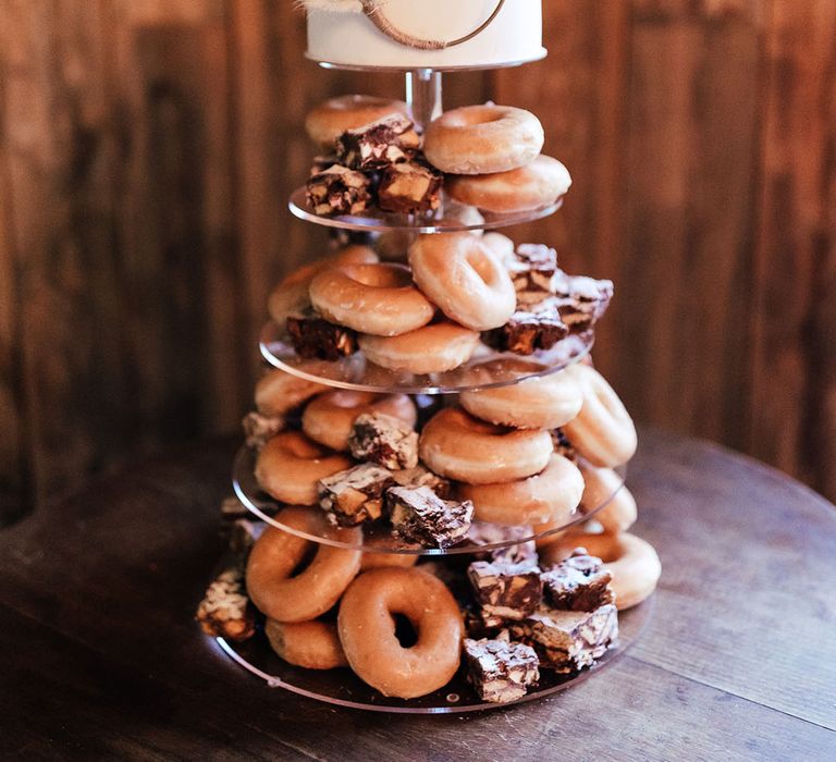 Glazed and iced doughnuts on acrylic cake stand with layer of white frosted cake at the top of the stand with pampas grass decoration