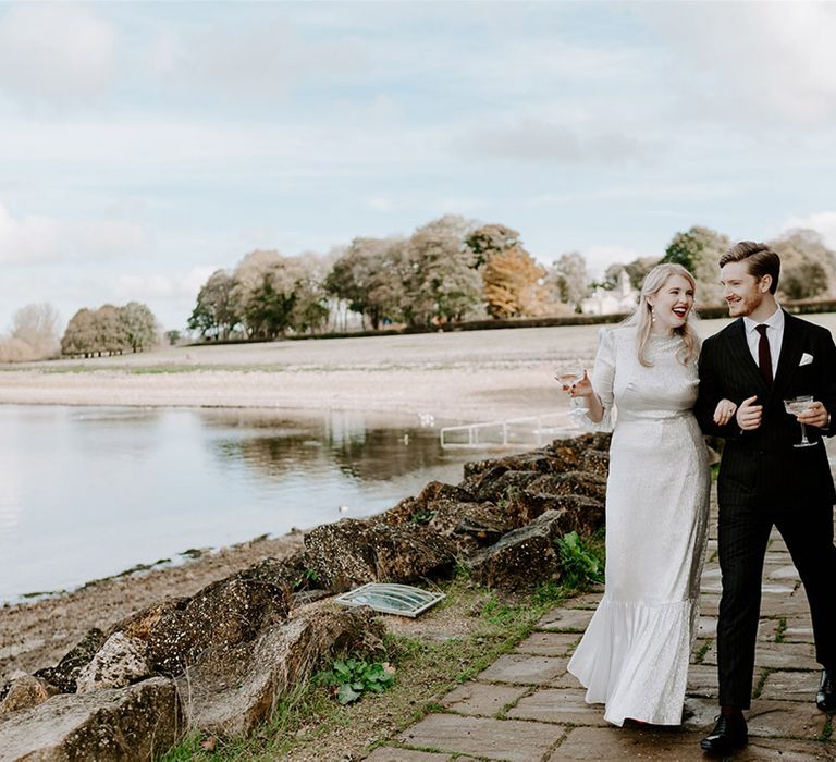 Bride in silver wedding dress walking with the groom in a pinstriped suit drinking champagne from coupe glasses 