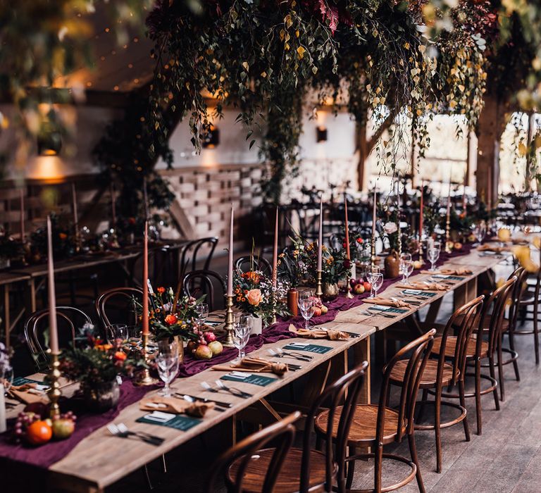 Orange and pink taper candles in gold candleholders with dark burgundy table runner with plenty of autumnal foliage and flowers 