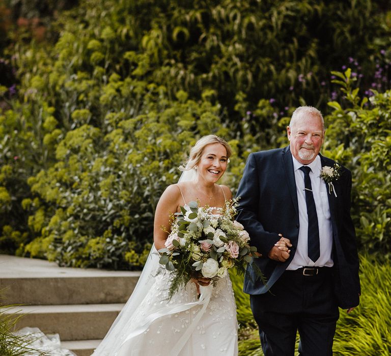 Bride in applique wedding dress by Alexandra Grecco walking to the ceremony with the father in a traditional black tie outfit 