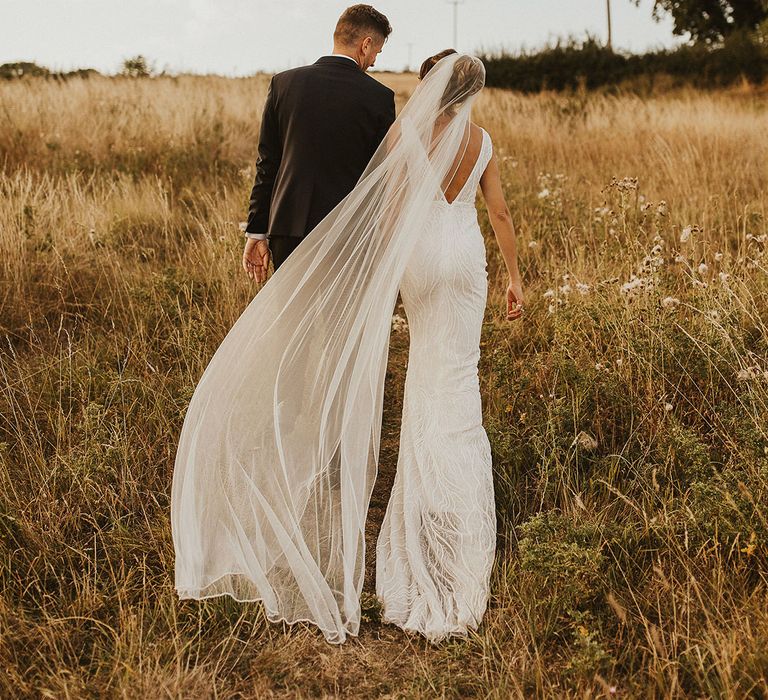 Bride in beaded wedding dress by Made With Love with her veil blowing in the wind walking with the groom in tuxedo 