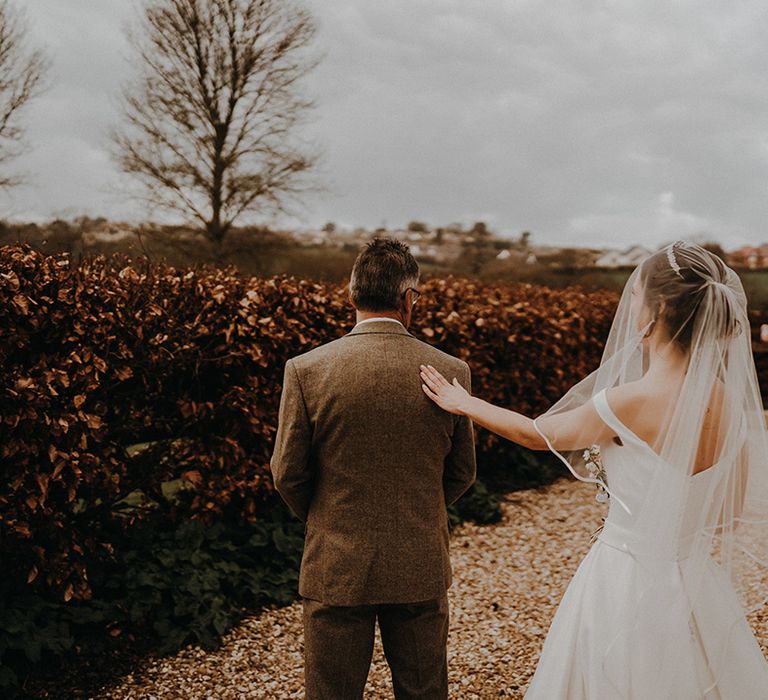 The bride wearing a traditional wedding dress with wedding veil approaches her father for an emotional first look moment 