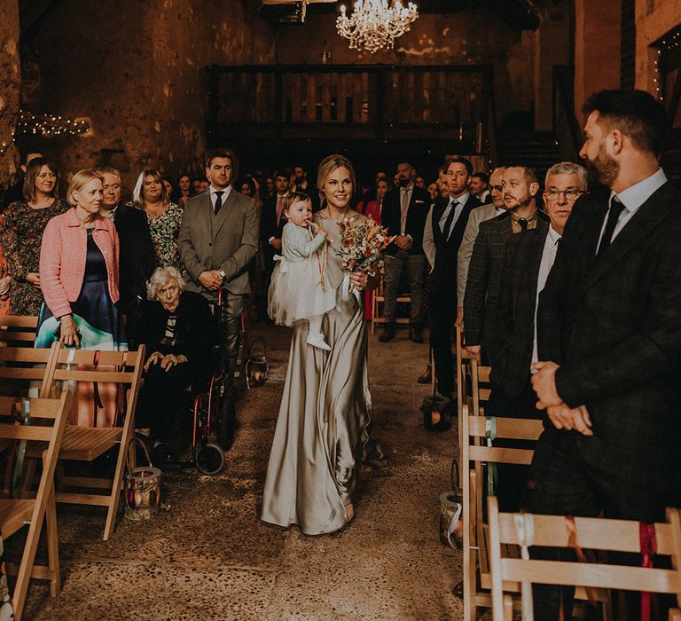 Maid of honour in satin sage green bridesmaid dress carrying a flower girl in a white dress down the aisle at barn wedding venue 