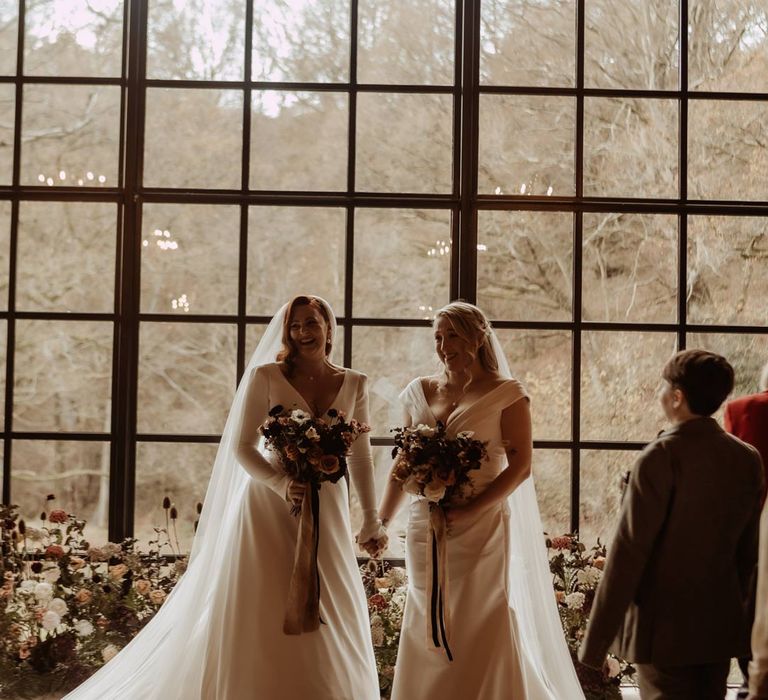 Brides in complimenting deep v neck wedding dresses standing by the window at Hidden River Cabins at LGBTQIA wedding 