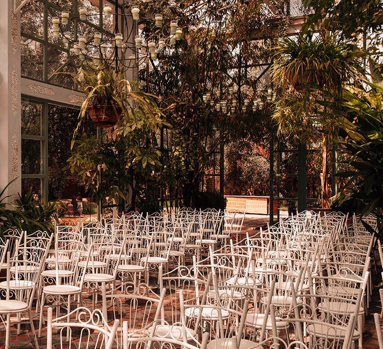 White chairs and botanical plants and foliage decorate the inside of the glass house wedding venue