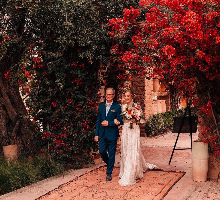 Father of the bride in blue suit and colourful tie walking the bride in modern lace bridal two piece down the aisle to the wedding ceremony 