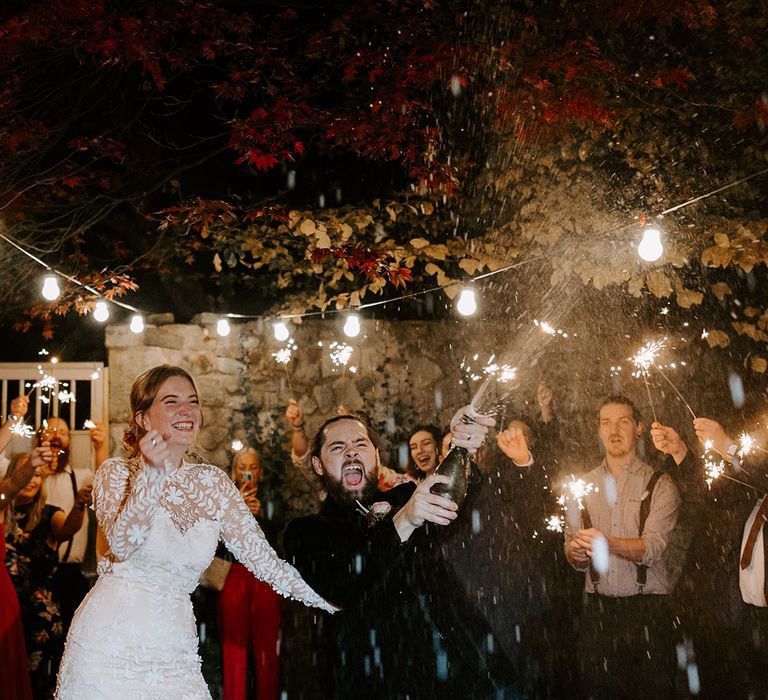 The groom in a black velvet suit pops champagne to celebrate the wedding with the bride and wedding guests 