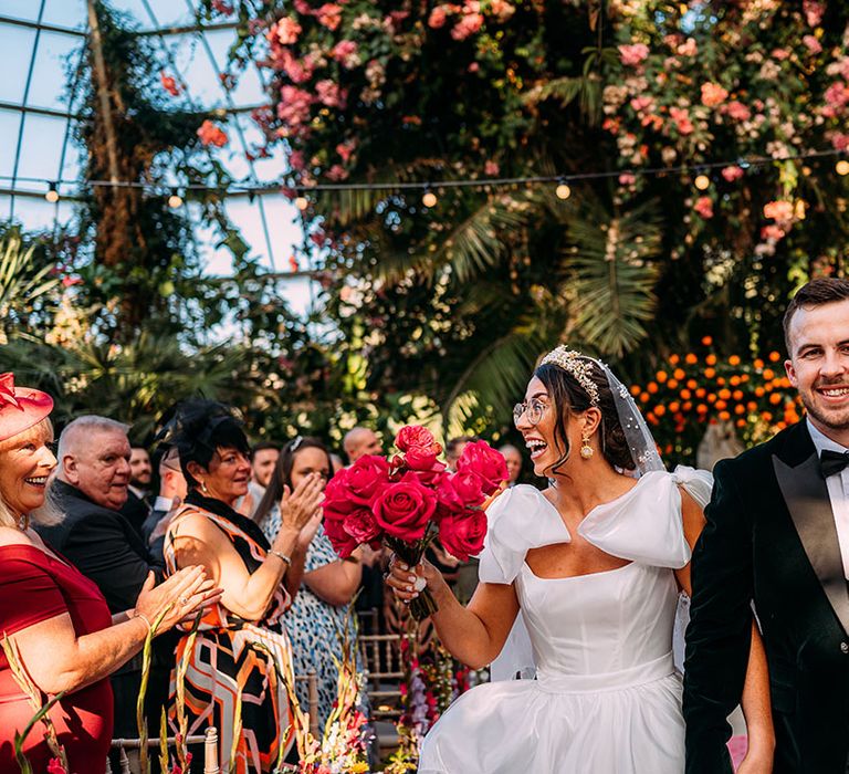 Bride wearing gold crown and statement Emma Beaumont wedding dress carrying a pink rose bouquet with groom in black tie 