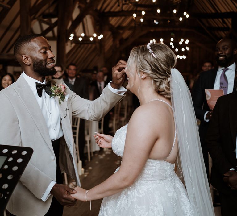 Black groom in wearing black bow tie and grey suit jacket wipes tears from the bride's eyes at the heartfelt ceremony 