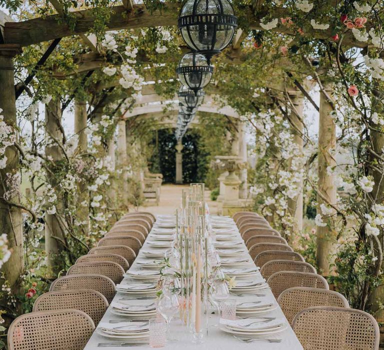 Blush pink tapered candles in glass candlestick covers on spring inspired wedding tablescape with white ruffled wedding table cloth, white peony, pink garden rose, foliage and blush florals wedding table centrepieces and simple crockery at Euridge Manor wedding