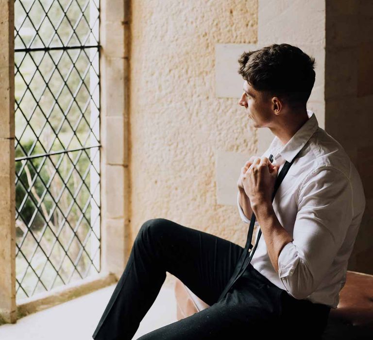 Groom sitting by a windowsill at Euridge Manor getting ready for wedding wearing crisp white shirt, black suit trousers and black tie around his neck 