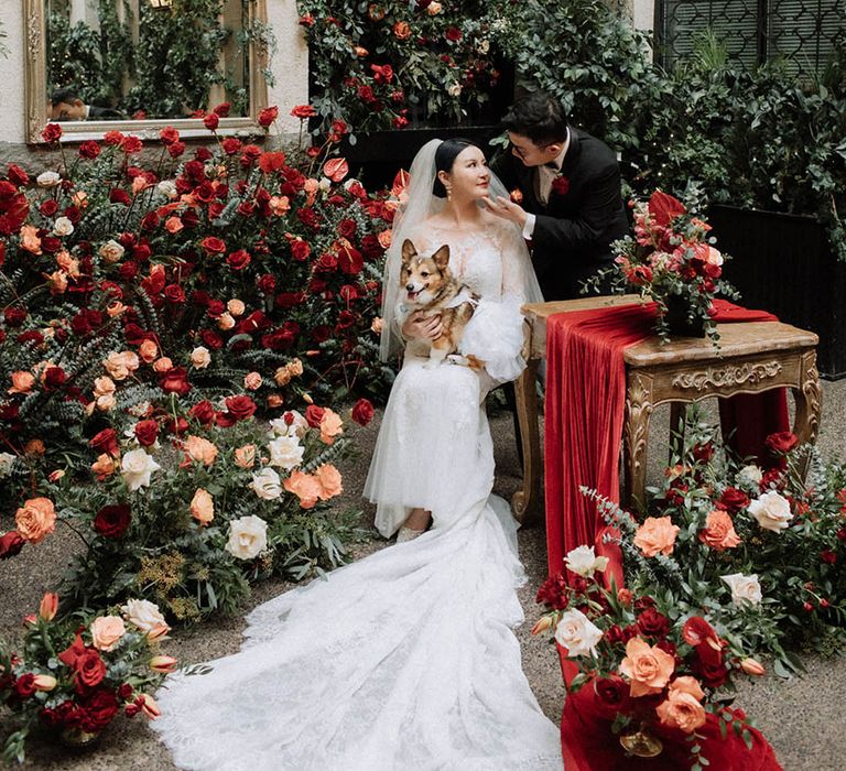 The bride and groom sit together with their pet dog surrounded by romantic red wedding flowers 