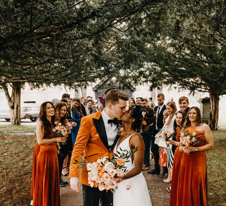 Groom in crushed burnt orange velvet grooms blazer, black bowtie and black suit trousers kissing bride in v-neck sleeveless wedding dress with 3D floral applique train with bridesmaids in one shoulder burnt orange bridesmaid dresses and groomsmen in classic black grooms suits at Brinsop Court wedding