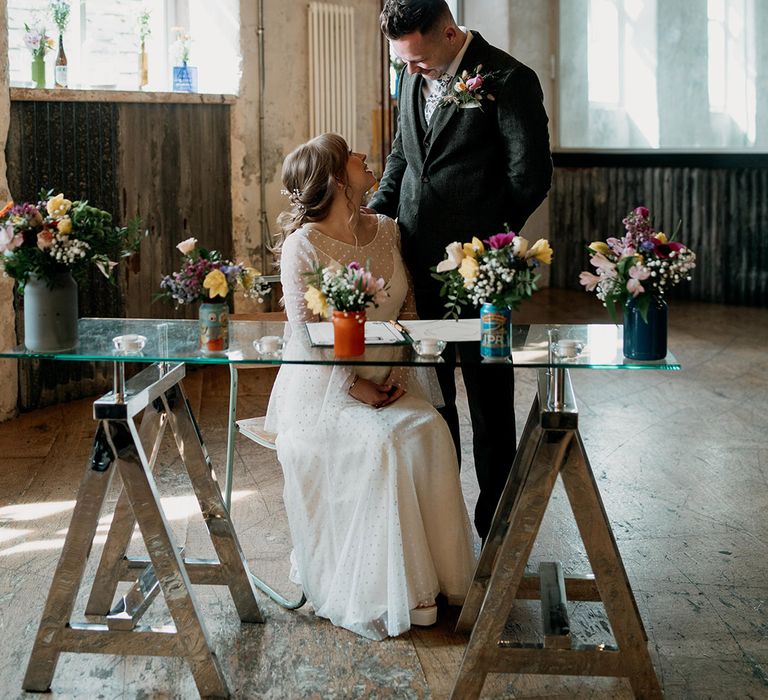 The bride and groom gaze lovingly into each other's eyes as they sit to sign the wedding register surrounded by wildflower wedding decor 