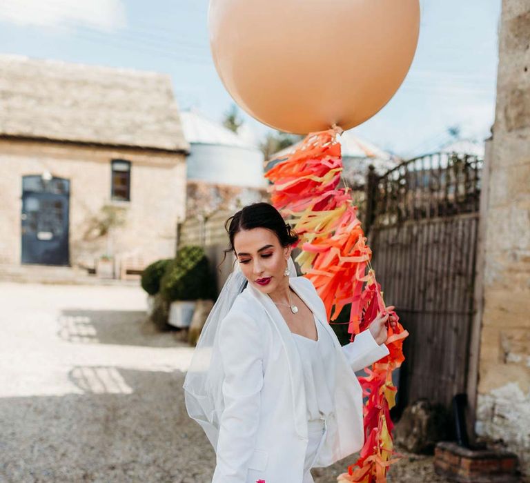 Bride in white bridal suit with closed toe bright green wedding heels holding large balloon decoration with wedding streamers attached outside of Merriscourt wedding venue