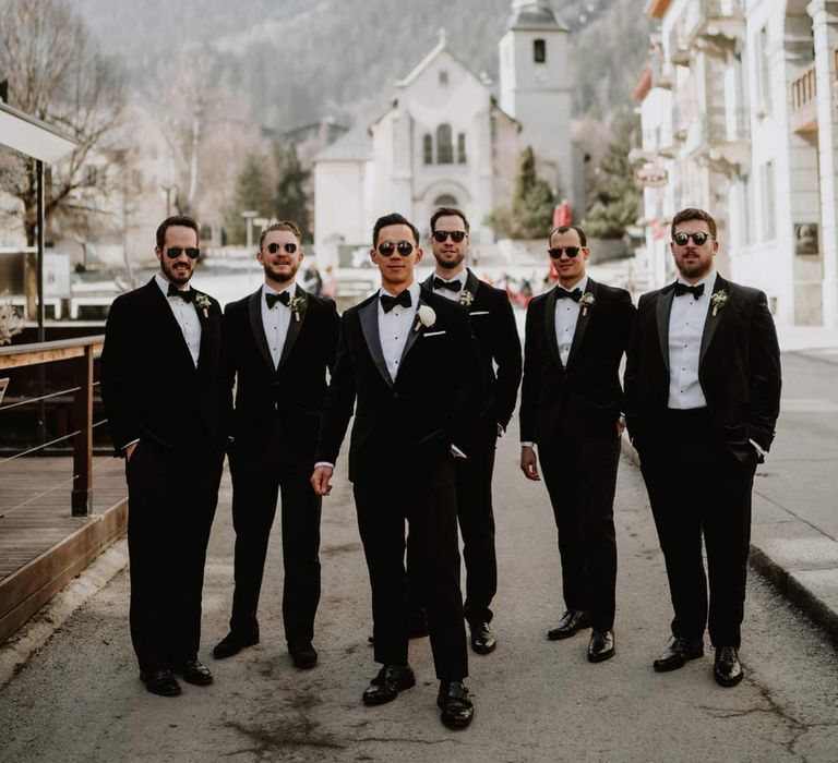 Groomsmen in matching classic black tuxedos, black bowtie sand white garden rose boutonnieres standing with the French Alps in the background at French destination ski wedding