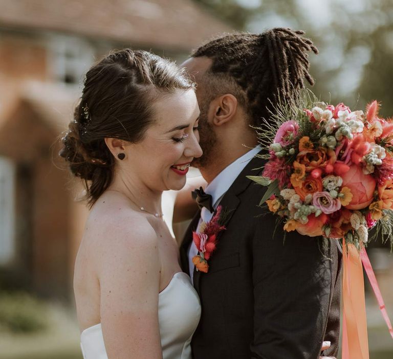 Groom in classic black tuxedo with bowtie and pink and orange wedding boutonniere embracing bride in strapless sweetheart neckline wedding dress holding pink and orange wedding bouquet 