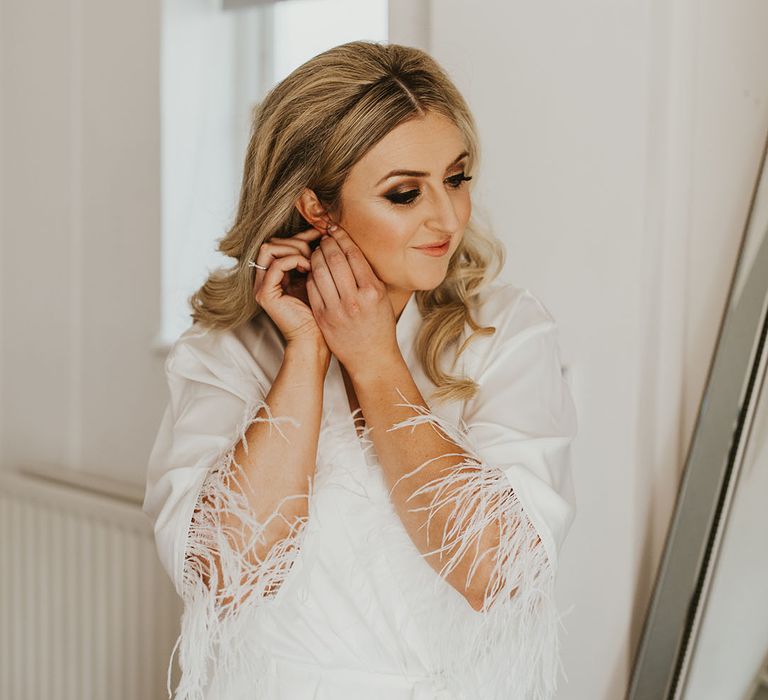 Bride in a white satin robe with ostrich feathers as she get ready for the wedding putting in her earrings 