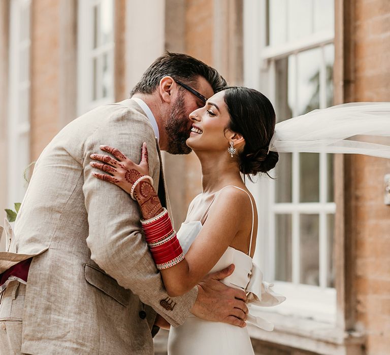 Bride wearing stunning wedding veil blowing in the wind kissing the groom in a cream three piece wedding suit 