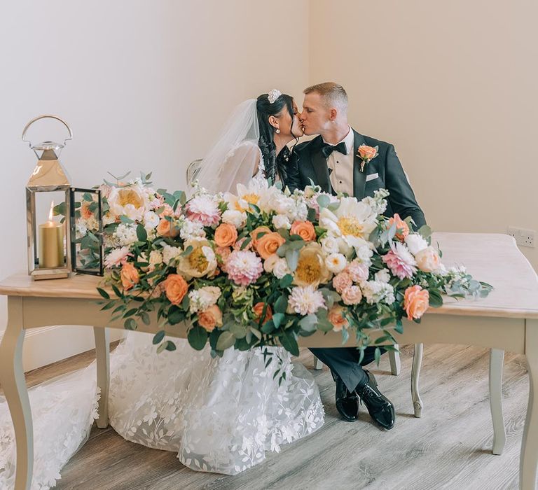 Bride and groom sit to sign their wedding register at a table decorated with seasonal flowers 