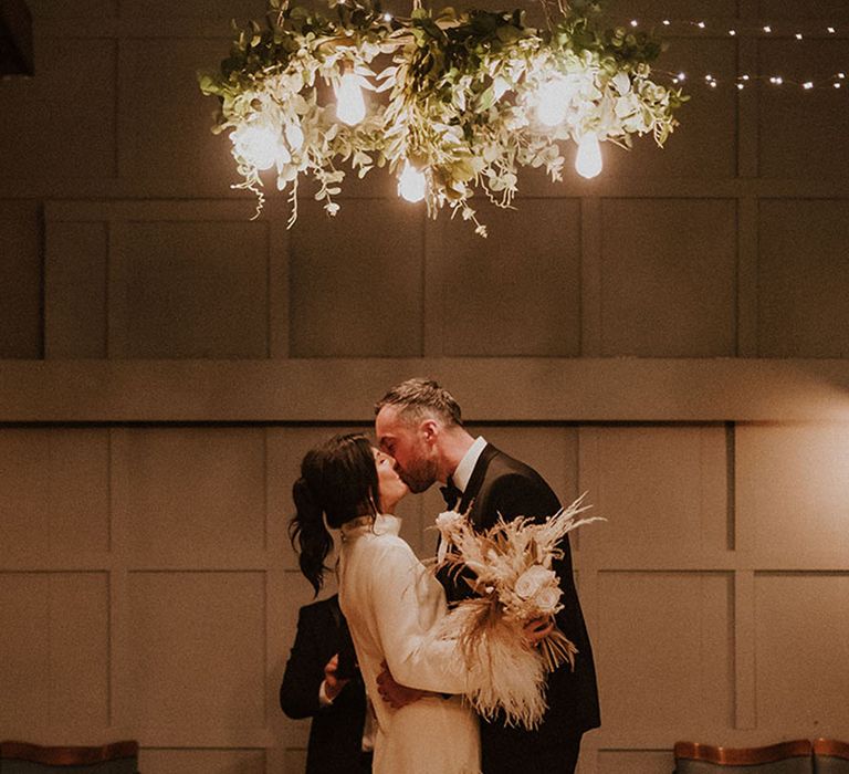 Bride carrying white neutral wedding bouquet with dried wedding flowers as she kisses the groom 